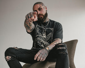 Man with tattoos sitting on a modern chair, pointing forward.