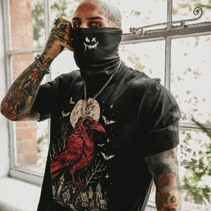 A tattooed man wearing a black short-sleeve graphic t-shirt with a red raven design and a black neck gaiter mask featuring a white skull graphic, standing by a window.