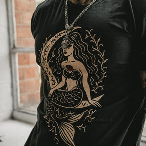 A close-up of a man wearing a black t-shirt with a large beige mermaid graphic print on the front, styled with a silver chain necklace.
