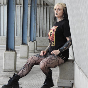 Person wearing a black t-shirt with a colorful design, fishnet stockings, and black boots sitting on a concrete ledge.