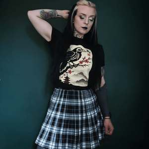 Person wearing a black t-shirt with a graphic design and a plaid skirt against a dark background