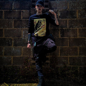 Person wearing a black t-shirt with a graphic design, standing against a brick wall with a bright light source in the background.