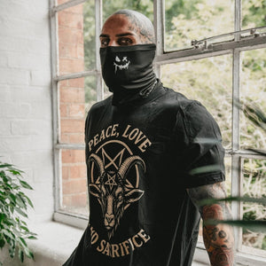 A man wearing a black t-shirt by inked 13 with a beige goat head graphic and the words 'PEACE, LOVE, AND SARIFICE' on the front, paired with a matching black face covering, leaning against a window in a bright room.