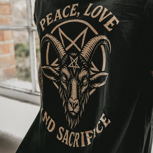 A person wearing a black long-sleeve t-shirt with a large beige graphic of a goat head inside a pentagram circle, with the words 'PEACE, LOVE' and 'AND SACRIFICE' printed around it. The person is also wearing a black neck gaiter with a white skull-and-crossbones logo.